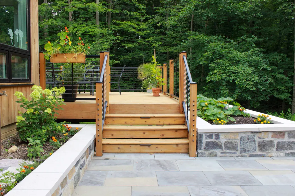 Wide wood stairs from deck to stone patio with raised stone beds, planting, railings, and wooded edge.